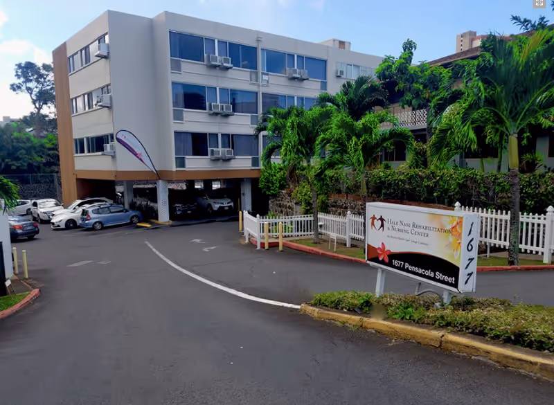Exterior view of Hale Nani Rehabilitation & Nursing Center showing a multi-story building with multiple windows and air conditioning units. There is a driveway leading to a covered parking area with several parked cars. The foreground features a sign with the facility's name and address, surrounded by palm trees and greenery.