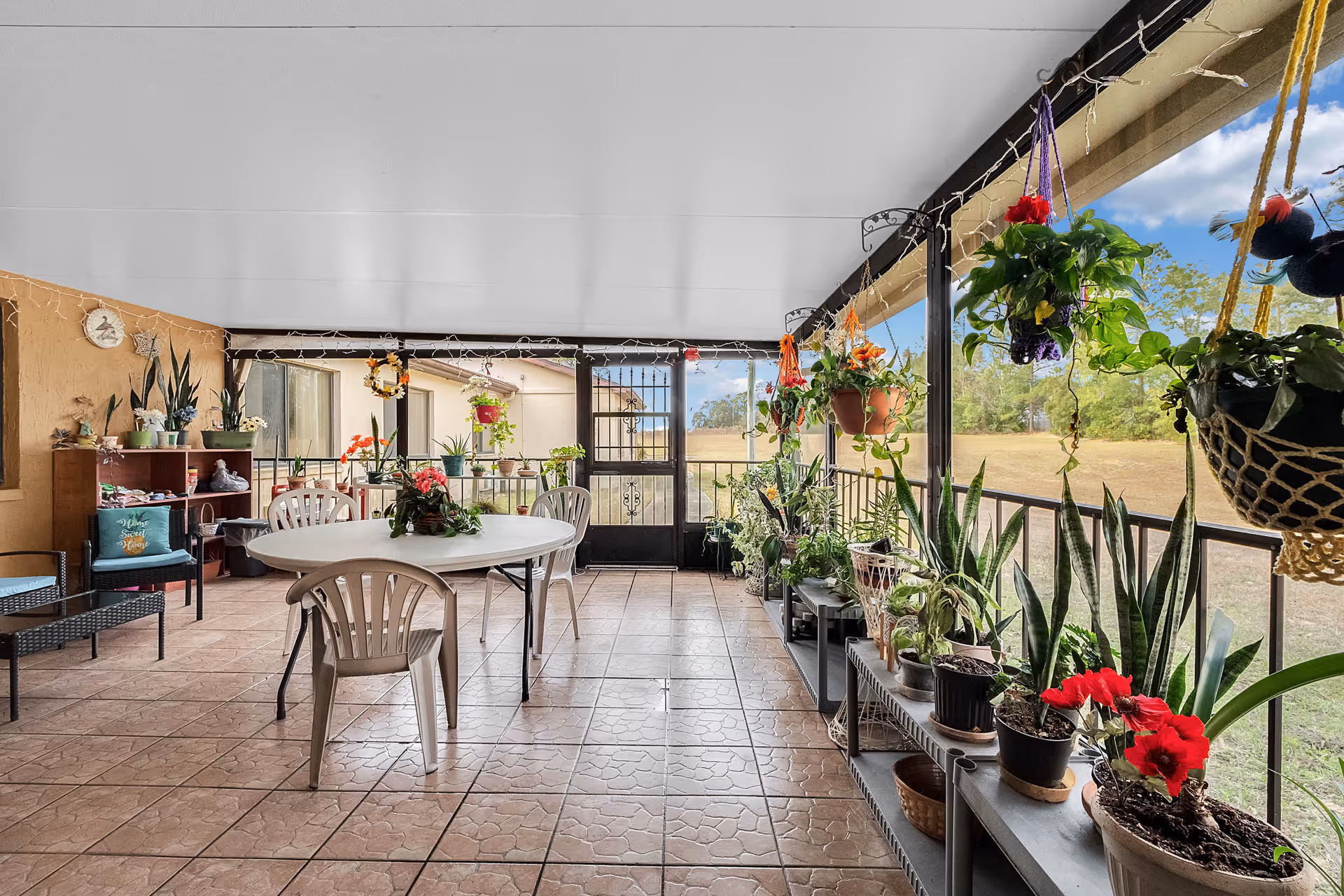 A covered patio area with tiled flooring featuring a white oval table surrounded by plastic chairs. The space is decorated with numerous potted plants on shelves and hanging from the ceiling. There is a black metal door leading outside to a grassy field with trees in the background. The patio has string lights hanging along the ceiling edges and some cushioned seating with a decorative pillow on the left side.