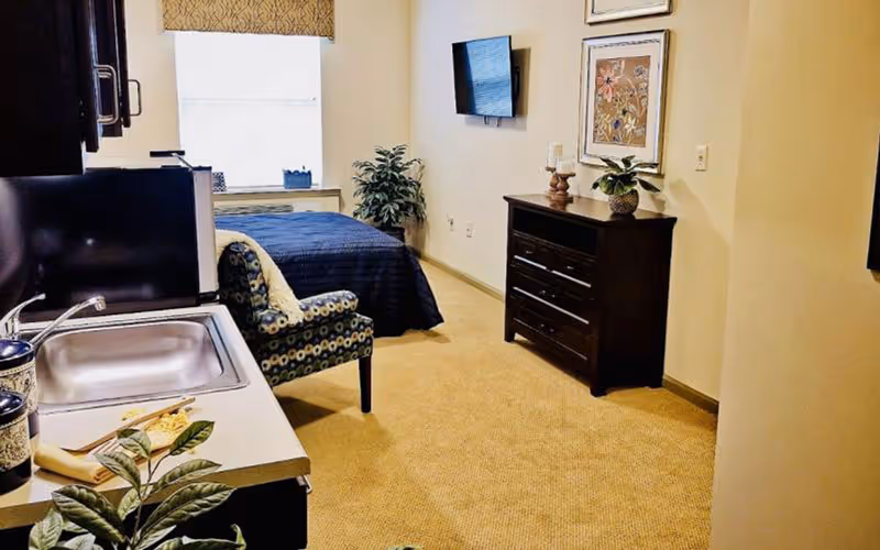 Interior view of a senior living facility room at Oak Point of Carthage featuring a small kitchenette with a sink and microwave on the left, a patterned armchair, a bed with a blue bedspread near a window, a potted plant, a wall-mounted TV, and a dark wooden dresser with decorative items and framed artwork above it.