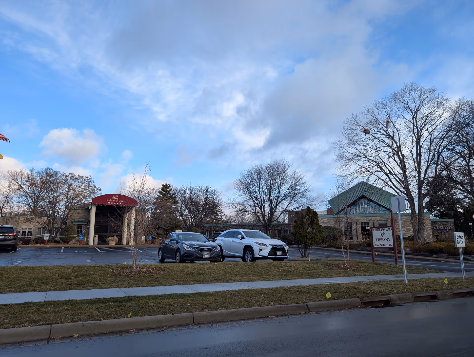 Exterior view of Heritage Of Edina II facility showing the building entrance with a red awning, parked cars in front, leafless trees, and a partly cloudy sky.