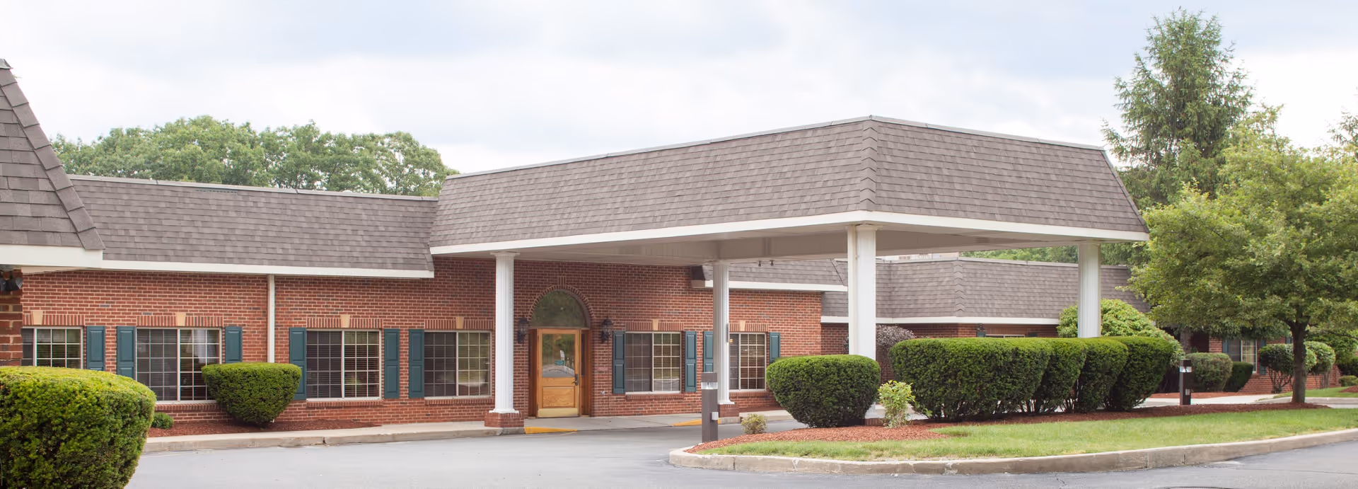 Brick single-story senior living facility entrance with a covered porte-cochere, trimmed shrubs, and a driveway.