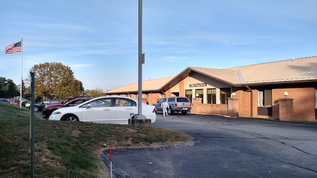 Single-story brick senior living facility entrance with several parked cars and an American flag on a flagpole.