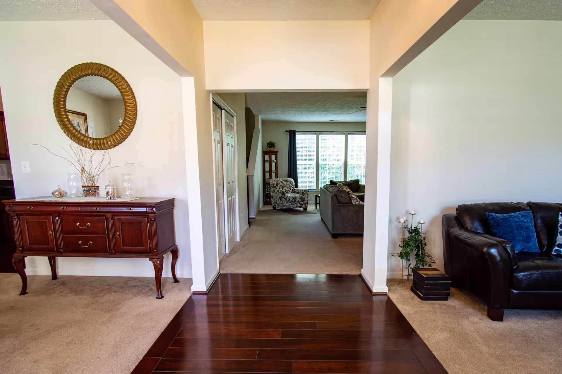 Interior view of a residential living space with a dark wooden floor leading to a carpeted living room area. On the left, there is a wooden sideboard with decorative items and a round mirror above it. On the right, there is a dark leather sofa with blue and patterned cushions, and a small plant stand with candles and greenery. The living room in the background has a patterned armchair, a gray sofa, and large windows with white blinds letting in natural light.