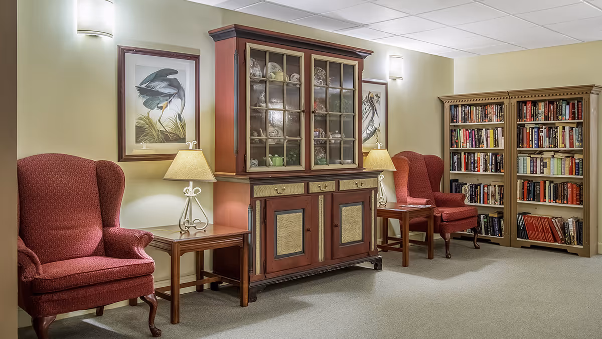 A cozy interior room with two red upholstered wingback chairs, two wooden side tables each with a lamp, a large wooden cabinet with glass doors displaying decorative items, two framed bird prints on the wall, and two bookshelves filled with books.