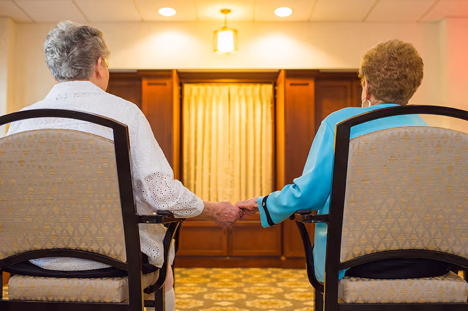 Two elderly women sitting on chairs facing a wooden cabinet with a curtain, holding hands in a warmly lit room.