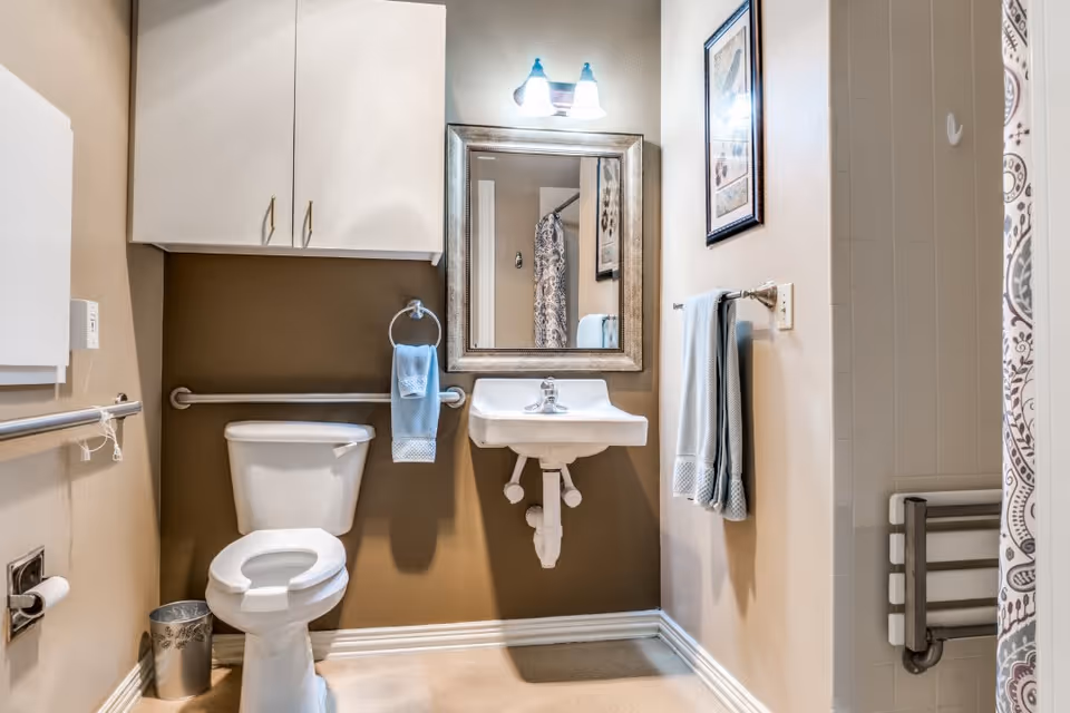 A clean and accessible bathroom with a white toilet, a wall-mounted sink with a silver faucet, a large framed mirror above the sink, and a shower area with a patterned curtain. There are light blue towels hanging on towel racks and a small trash bin next to the toilet. The walls are painted in beige and brown tones, and there are safety grab bars installed near the toilet and shower.