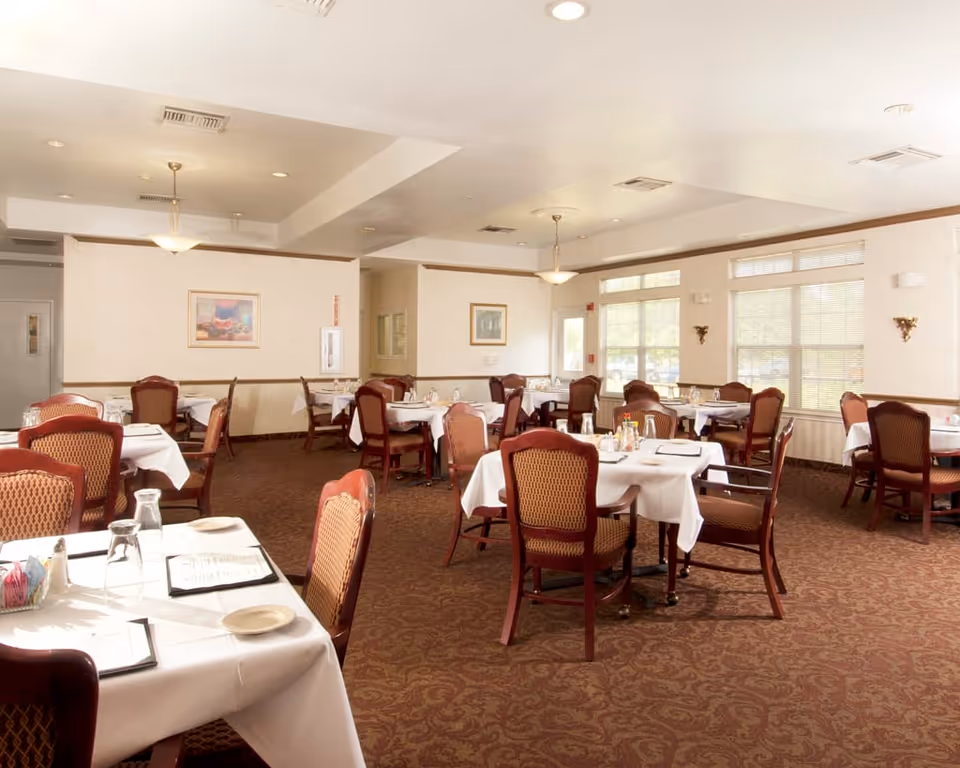 A bright dining room with multiple tables covered in white tablecloths, each set with plates, glasses, and menus. The room has patterned carpet, cushioned wooden chairs, large windows letting in natural light, and framed artwork on the walls.