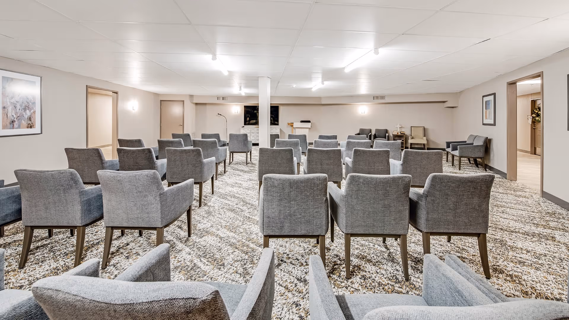 A meeting or activity room with multiple rows of gray upholstered chairs arranged facing a podium and a TV mounted on the wall. The room has beige walls, a patterned carpet, and framed artwork on the walls. There are doorways on both sides of the room leading to other areas.