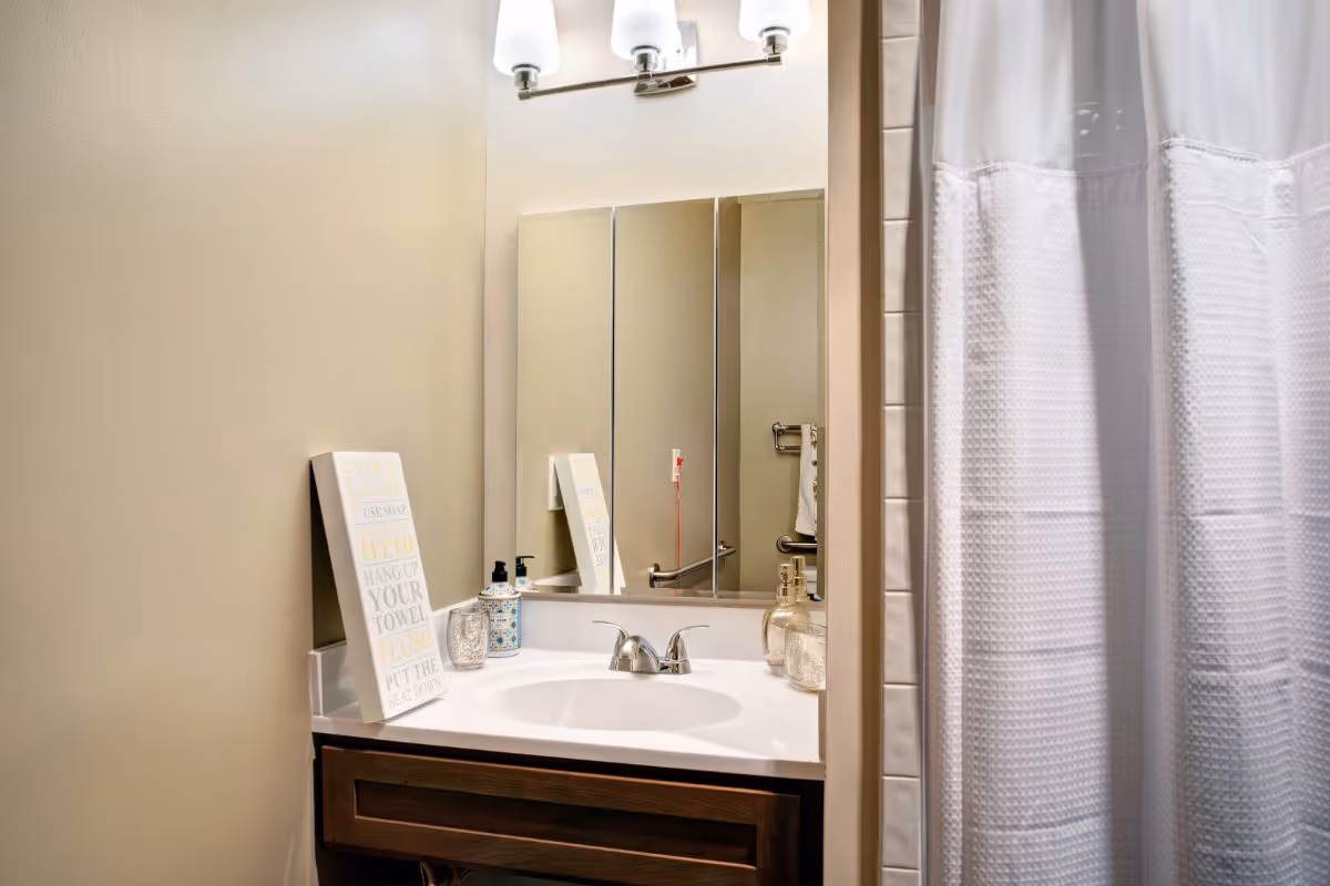 Small bathroom with a sink vanity, mirror and light fixture, toiletries on the counter, and a white shower curtain.