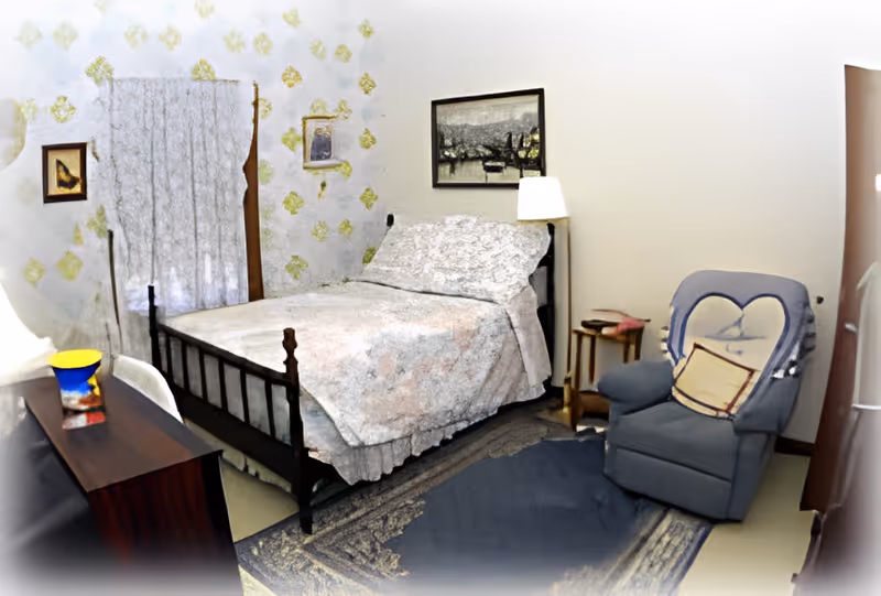 A cozy bedroom with a bed covered in a floral bedspread, a blue armchair with a heart-shaped pillow, a wooden nightstand with a lamp, and framed artwork on the walls. There is a window with white curtains and a wooden dresser with a colorful vase on top.