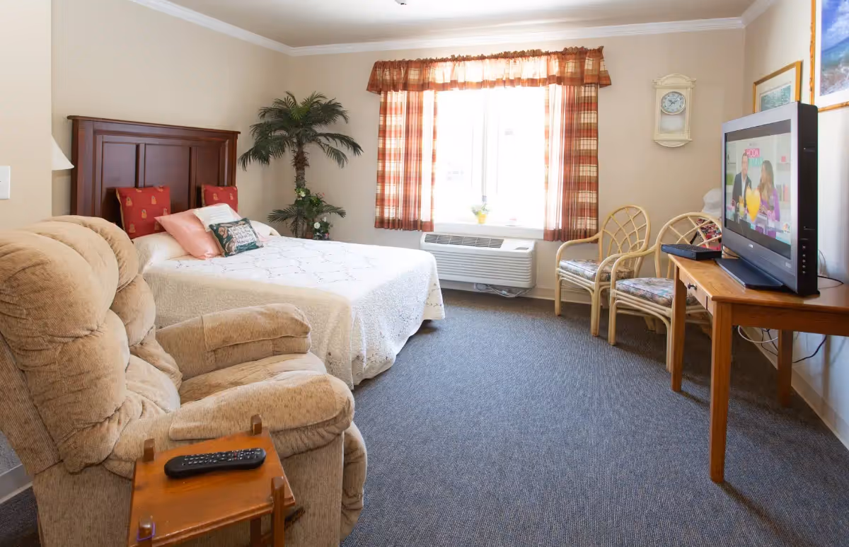 A cozy bedroom in an assisted living facility featuring a bed with a wooden headboard, multiple pillows, a beige recliner chair with a remote on a small wooden side table, two wicker chairs with cushions, a wooden table holding a flat-screen TV, a window with plaid curtains, a wall clock, and a potted plant in the corner.