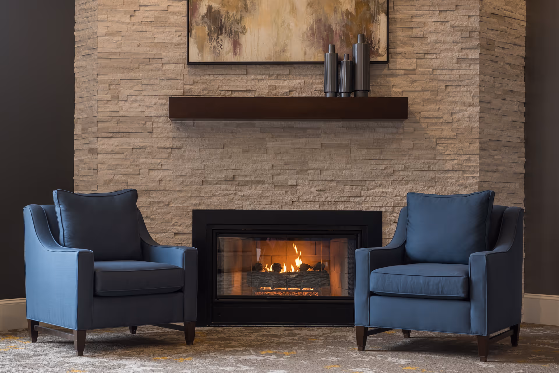 Two blue armchairs flanking a lit fireplace set in a textured stone wall with a floating shelf and artwork above.