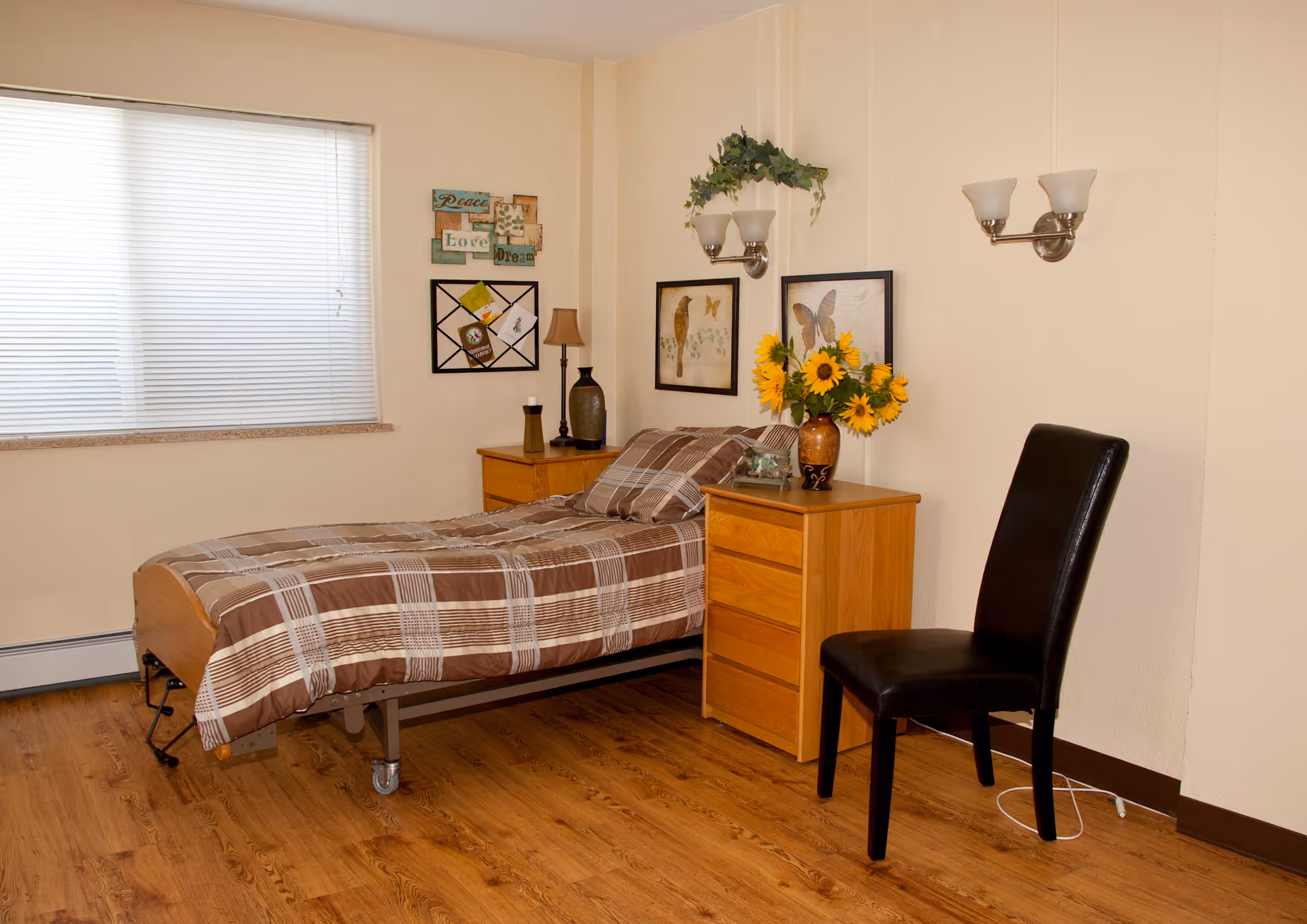 A tidy single-bedroom room with a twin bed, wooden bedside table and dresser, a black chair, sunflowers, and wall art.