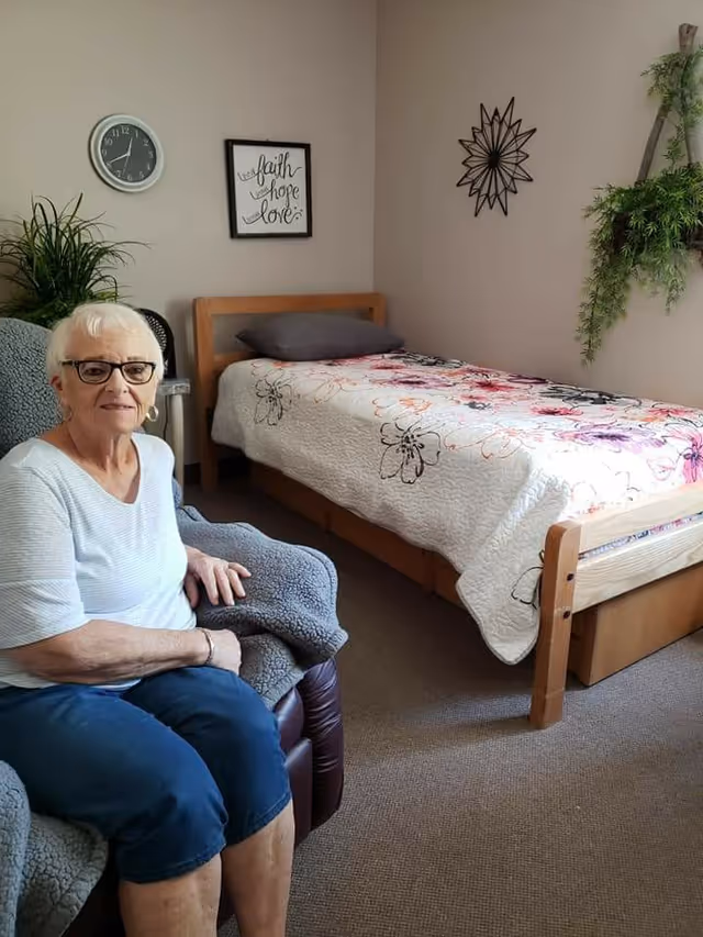 An elderly woman with short white hair and glasses sits on a cushioned chair in a cozy room. Behind her is a single bed with a floral quilt and a gray pillow. The room has beige walls decorated with a clock, a framed sign that reads 'faith hope love,' a metal starburst wall decoration, and a hanging green plant.