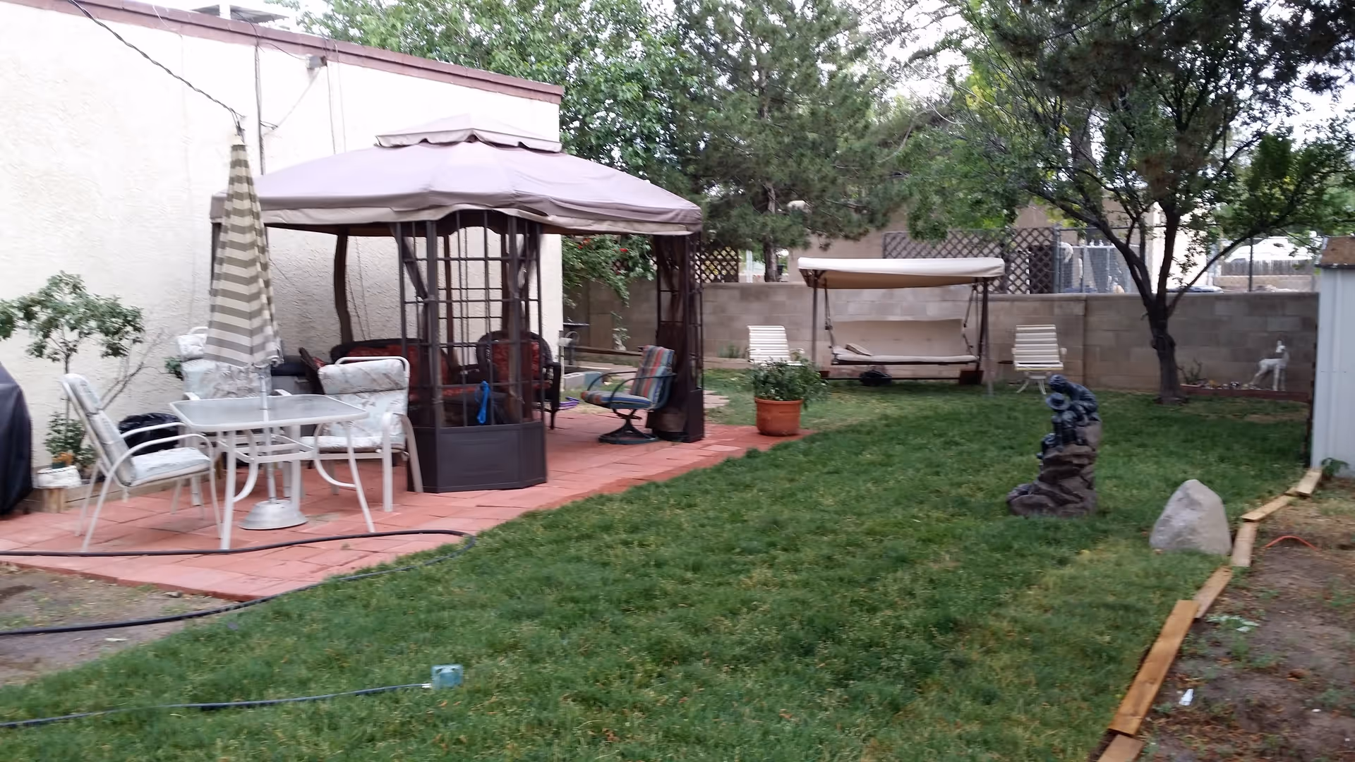 Outdoor garden area with green grass, a patio with a table and chairs under a striped umbrella, a gazebo with seating, a swing bench, trees, and decorative statues near a block wall fence.
