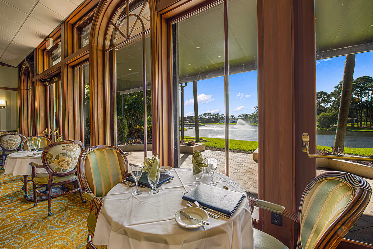 Dining area inside The Barclay at Sarasota with round tables covered in white tablecloths, set with plates, utensils, glasses, and folded napkins. The room features large wooden framed windows and doors offering a view of a patio, a lake with a fountain, and trees under a blue sky.