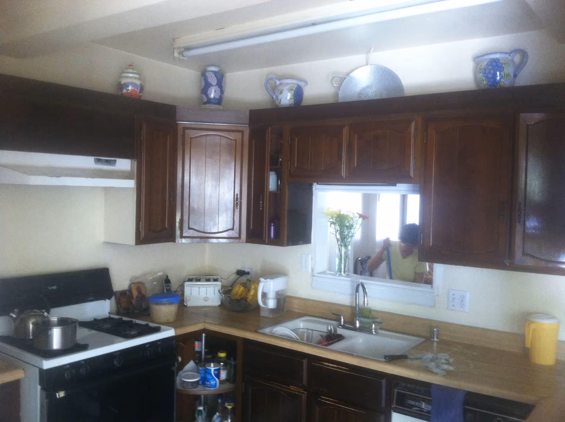 A kitchen with dark wooden cabinets, a white gas stove with a pot and kettle on it, a countertop with various items including a toaster, bananas, a water pitcher, and a sink with a faucet. Above the cabinets are decorative ceramic pitchers and a metal tray. There is a pass-through window with a vase of flowers and a person visible through it.