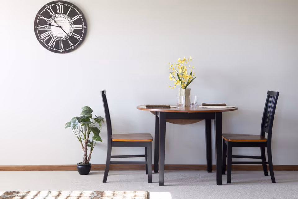 A small dining area with a round wooden table set for two with placemats, glasses, and a vase of yellow flowers. Two black wooden chairs with brown seats are placed on either side of the table. A potted green plant is on the floor to the left of the table, and a large black and white wall clock with Roman numerals hangs on the plain white wall behind.