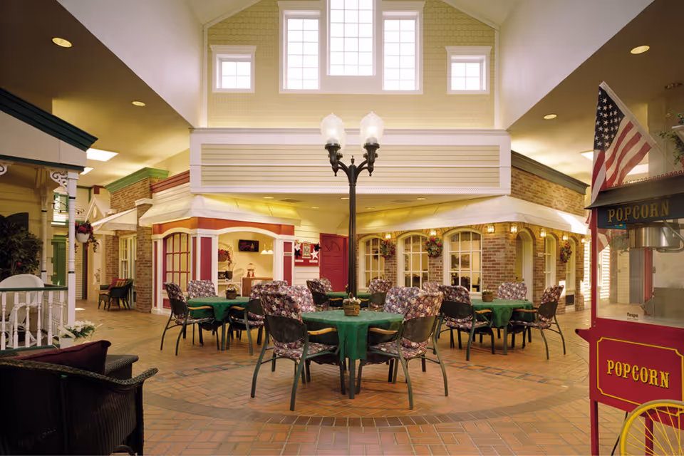 Indoor common area designed to look like a small town square with tables covered in green tablecloths and floral cushioned chairs arranged around them. There is a vintage-style popcorn machine on the right side and a streetlamp in the center. The walls have brick and siding facades with windows and flower baskets, and there are high windows letting in natural light.