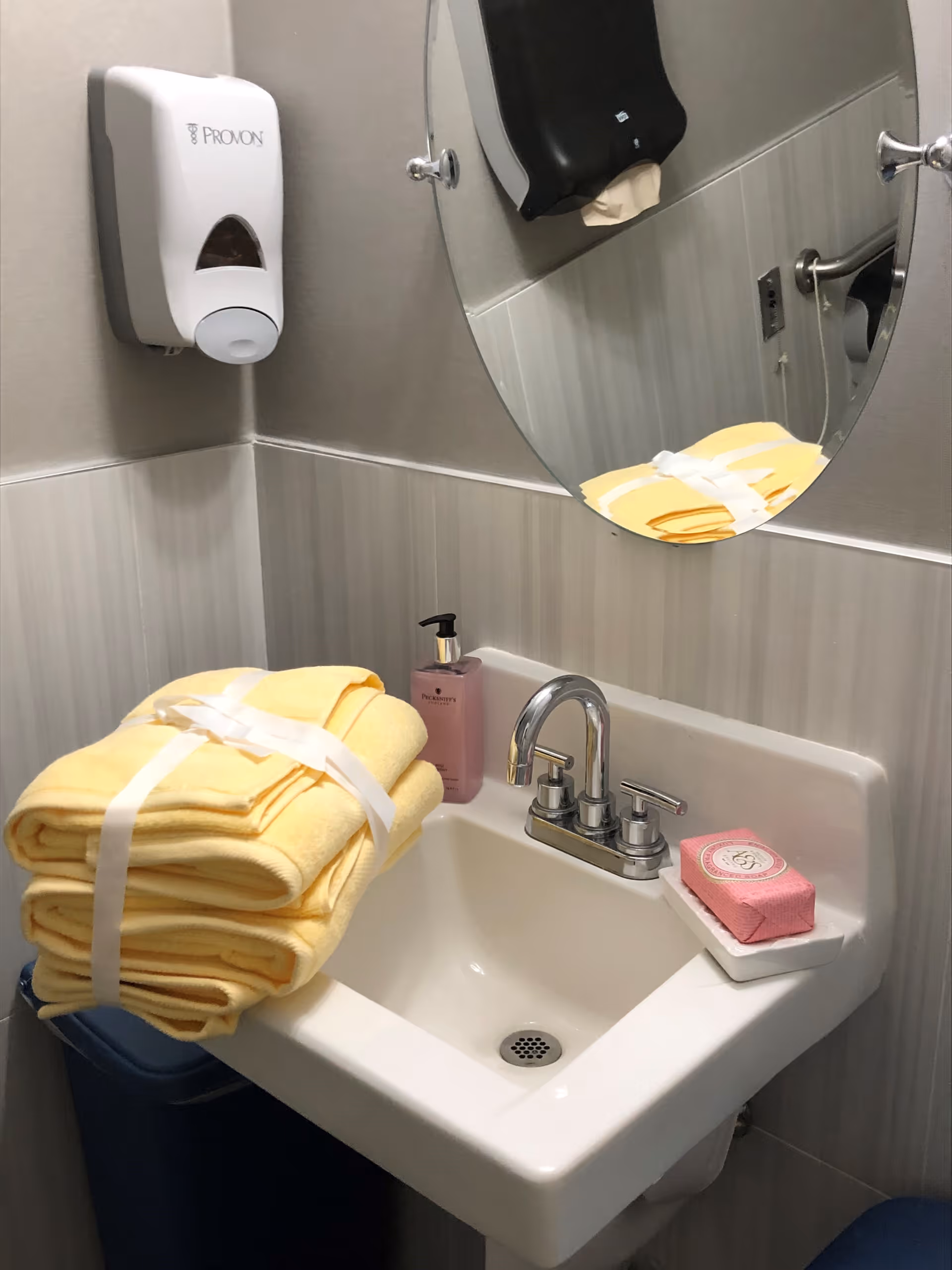 A small bathroom sink area with a stack of yellow towels tied with a white ribbon placed on the left side of the sink. There is a pink liquid soap dispenser and a pink bar of soap in a white soap dish on the right side of the sink. Above the sink is a round mirror and a wall-mounted soap dispenser on the left wall.