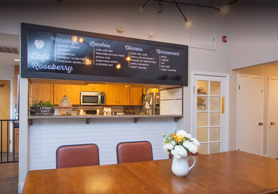 Interior view of a dining area with a wooden table and chairs. A white pitcher with flowers is placed on the table. In the background, there is a kitchen with wooden cabinets, a microwave, and a refrigerator. Above the kitchen counter, a large blackboard menu displays meal options for mornings, lunches, dinners, and amusement at Cafe Roseberry.