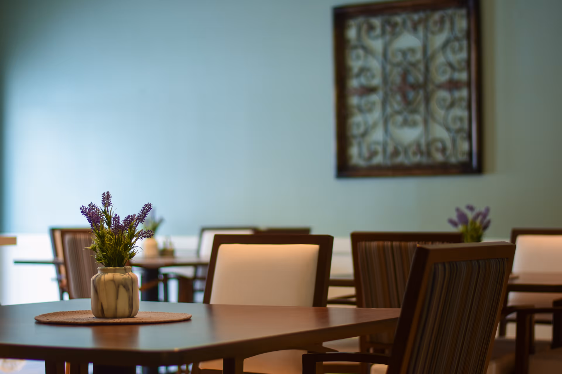 A dining area with wooden tables and chairs, each table decorated with a small vase containing purple flowers. The background shows a light blue wall with a framed decorative piece hanging on it.