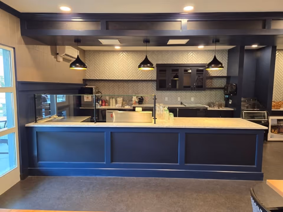 Interior view of a navy-blue serving counter and kitchen area with pendant lights and a glass sneeze guard.