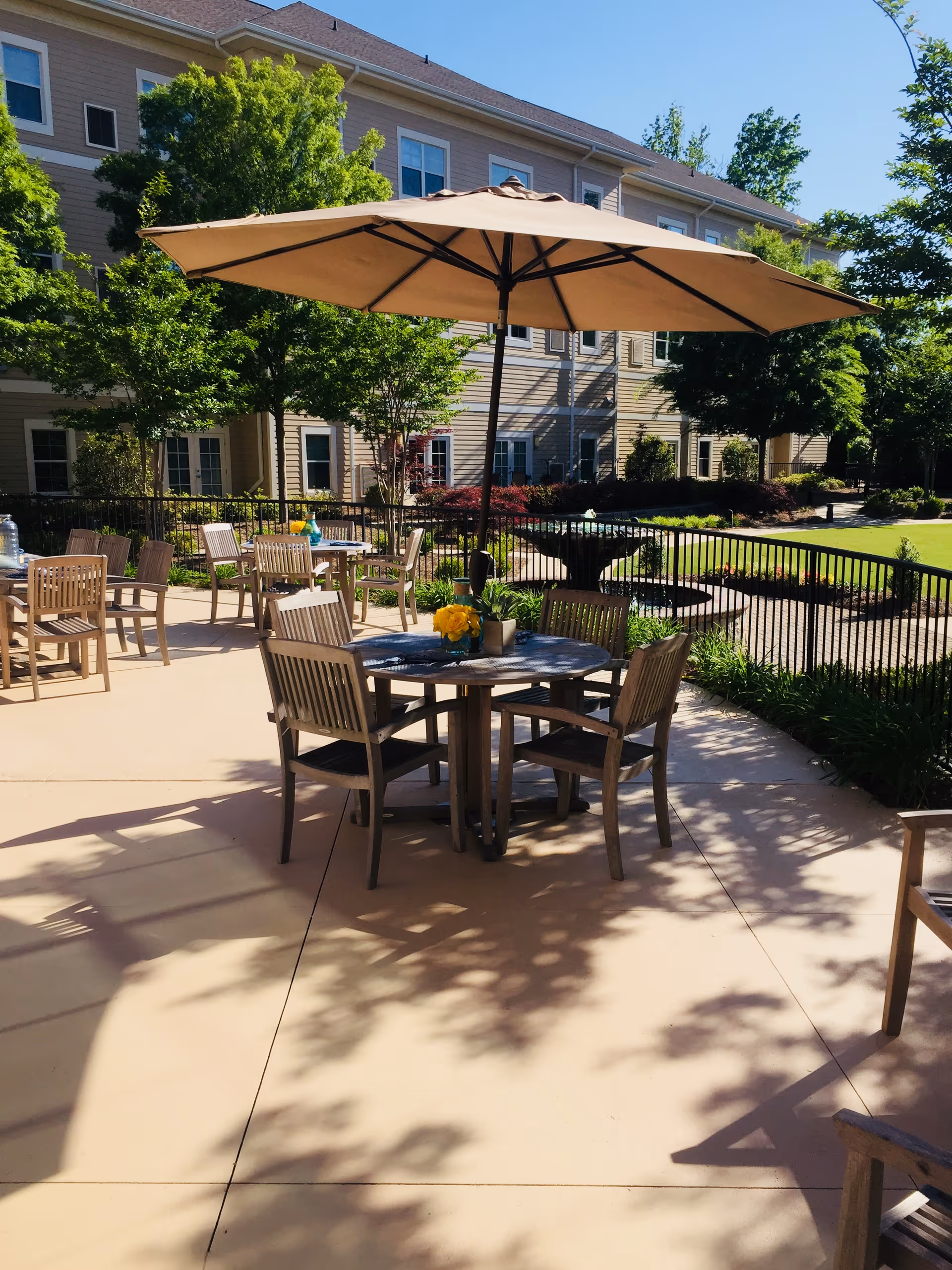 Outdoor patio area at Sterling Estates of East Cobb Retirement Community with wooden tables and chairs, large beige umbrellas providing shade, surrounded by greenery and a multi-story building in the background.