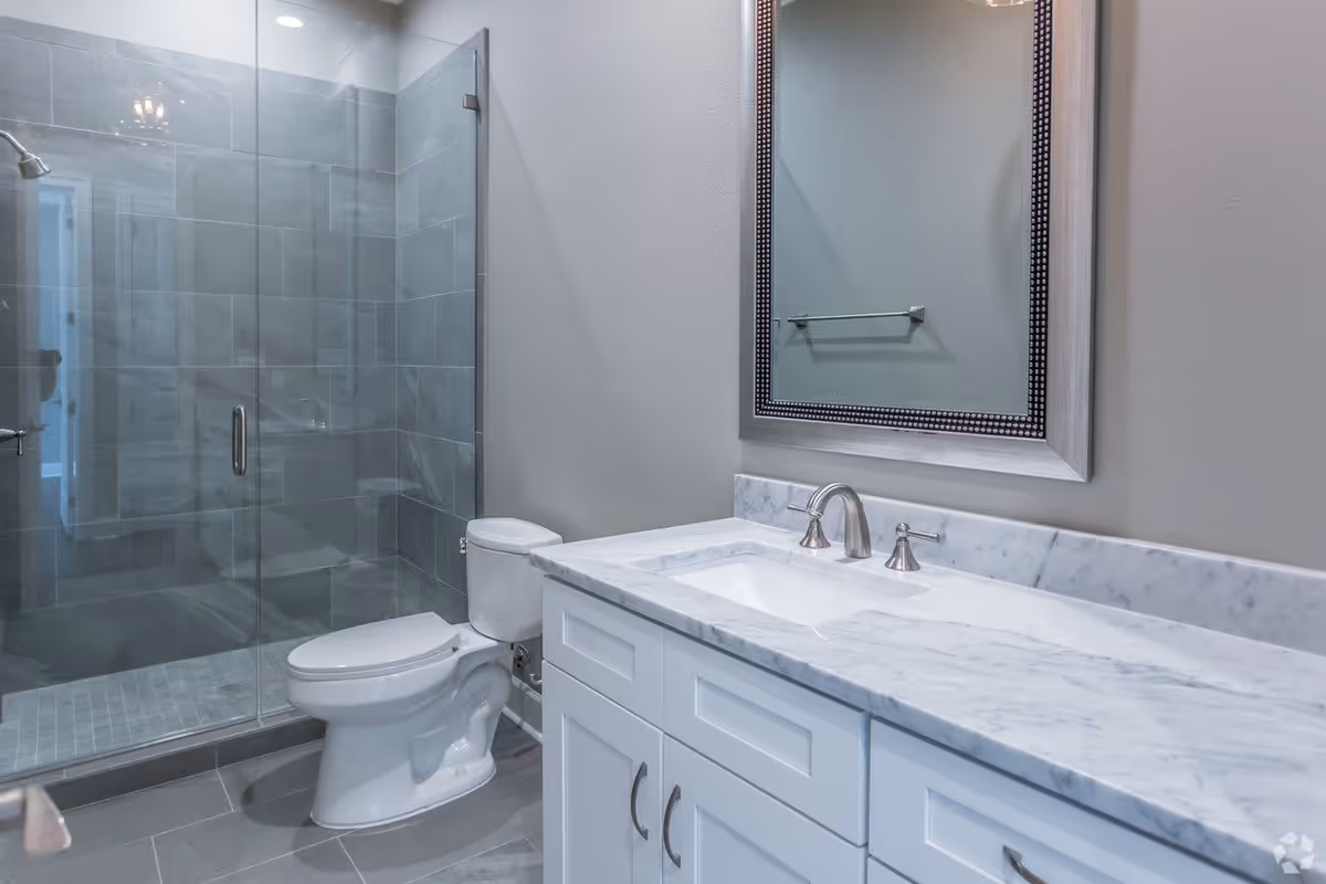 Clean modern bathroom with glass-enclosed shower, white toilet, and marble-topped vanity with mirror.