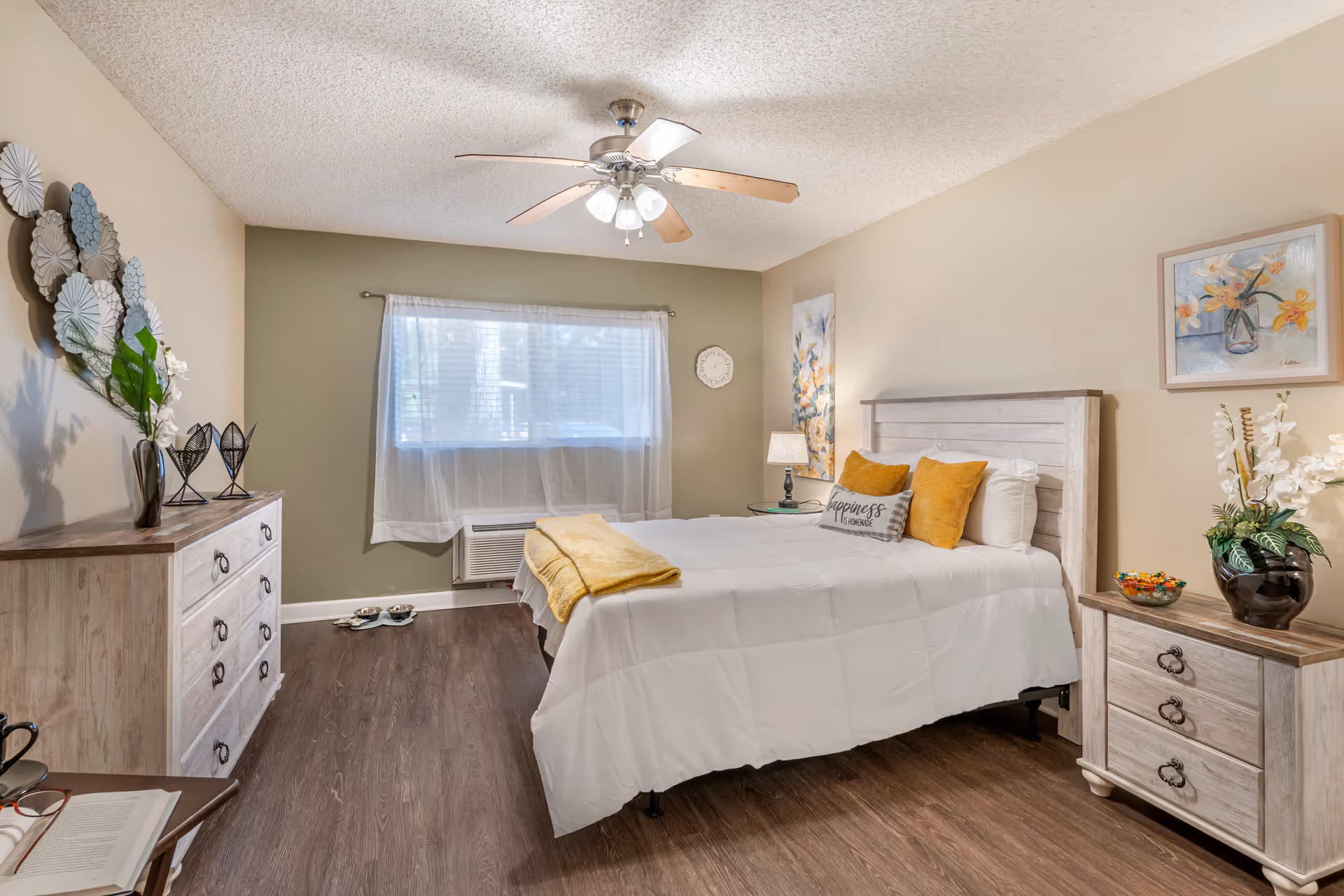 Bright furnished bedroom with a bed, nightstands, dresser, ceiling fan, and a window with sheer curtains.