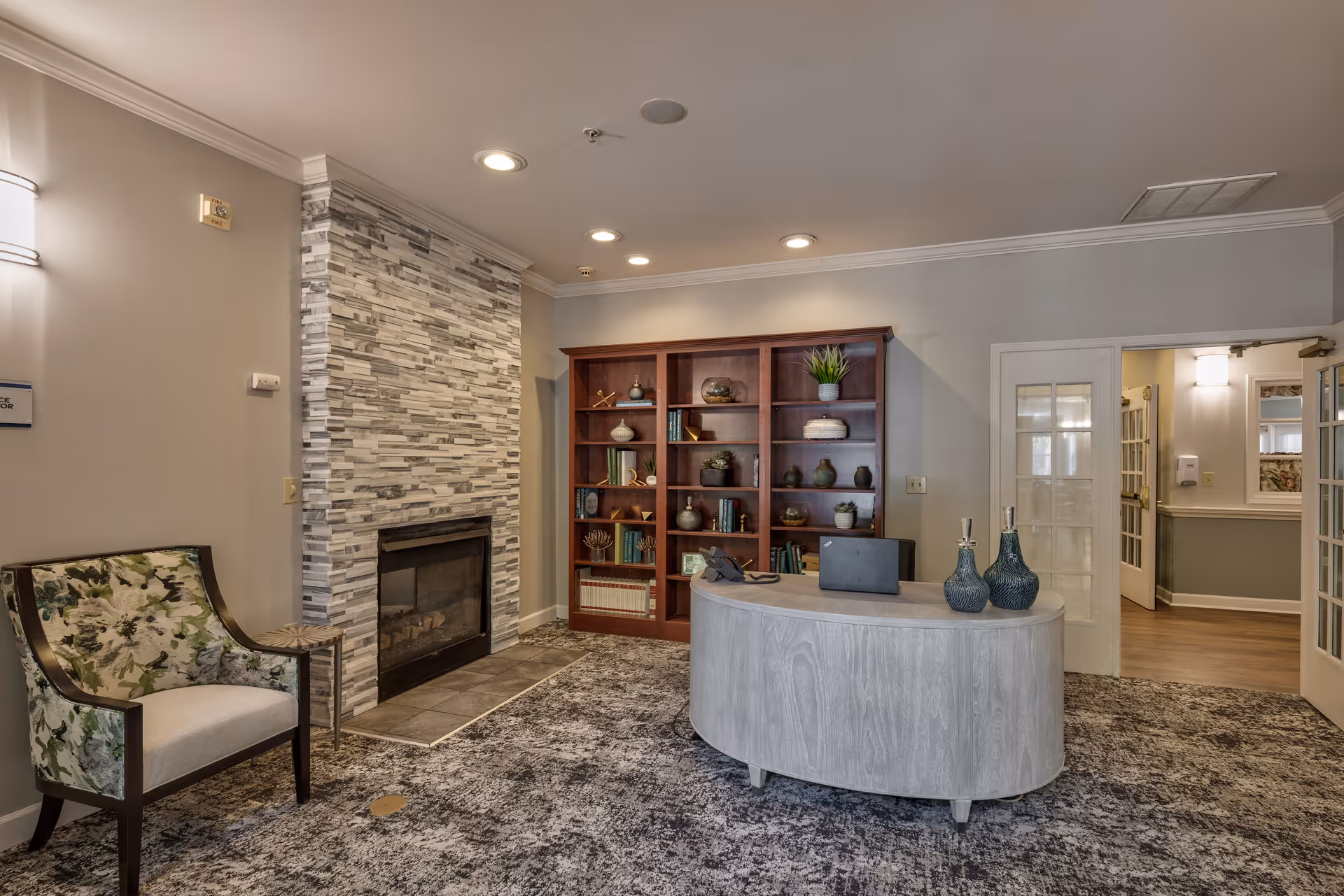 A cozy interior room featuring a stone fireplace with a tiled hearth, a wooden bookshelf filled with decorative items and books, a curved wooden reception desk with decorative vases, and a floral upholstered armchair next to a small side table. The room has recessed ceiling lights and an open doorway leading to another area.