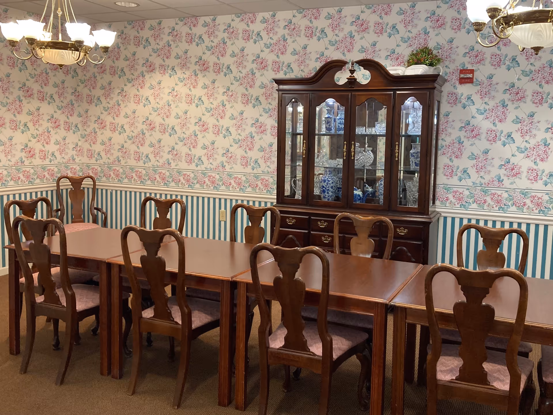 A dining room with a long wooden table surrounded by wooden chairs with cushioned seats. The walls are decorated with floral and striped wallpaper, and a wooden china cabinet filled with glassware and blue and white ceramics is against the wall. Two chandeliers with multiple lights hang from the ceiling.