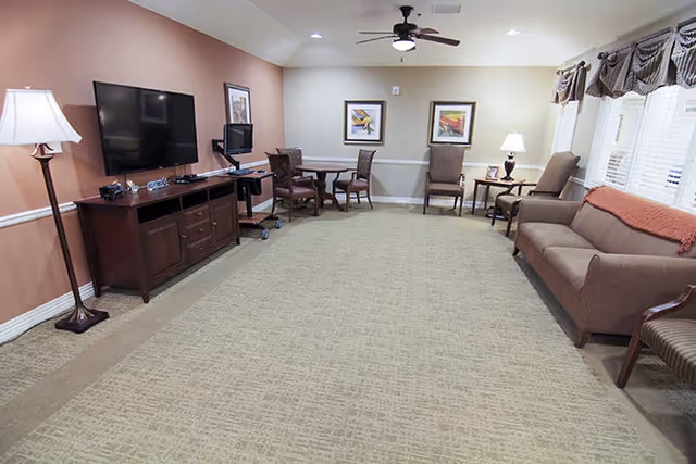 A spacious common living room with a sofa, armchairs, a wall-mounted TV on a wooden console, small tables, and a carpeted floor.