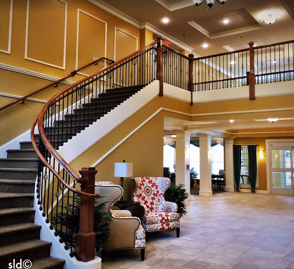 Interior view of a senior living facility lobby featuring a carpeted staircase with wooden handrails and black iron balusters. Two patterned armchairs and a small table with a lamp are placed near the base of the stairs. The walls are painted yellow with white trim, and the floor is tiled. The area is well-lit with ceiling lights and has large windows with curtains in the background.
