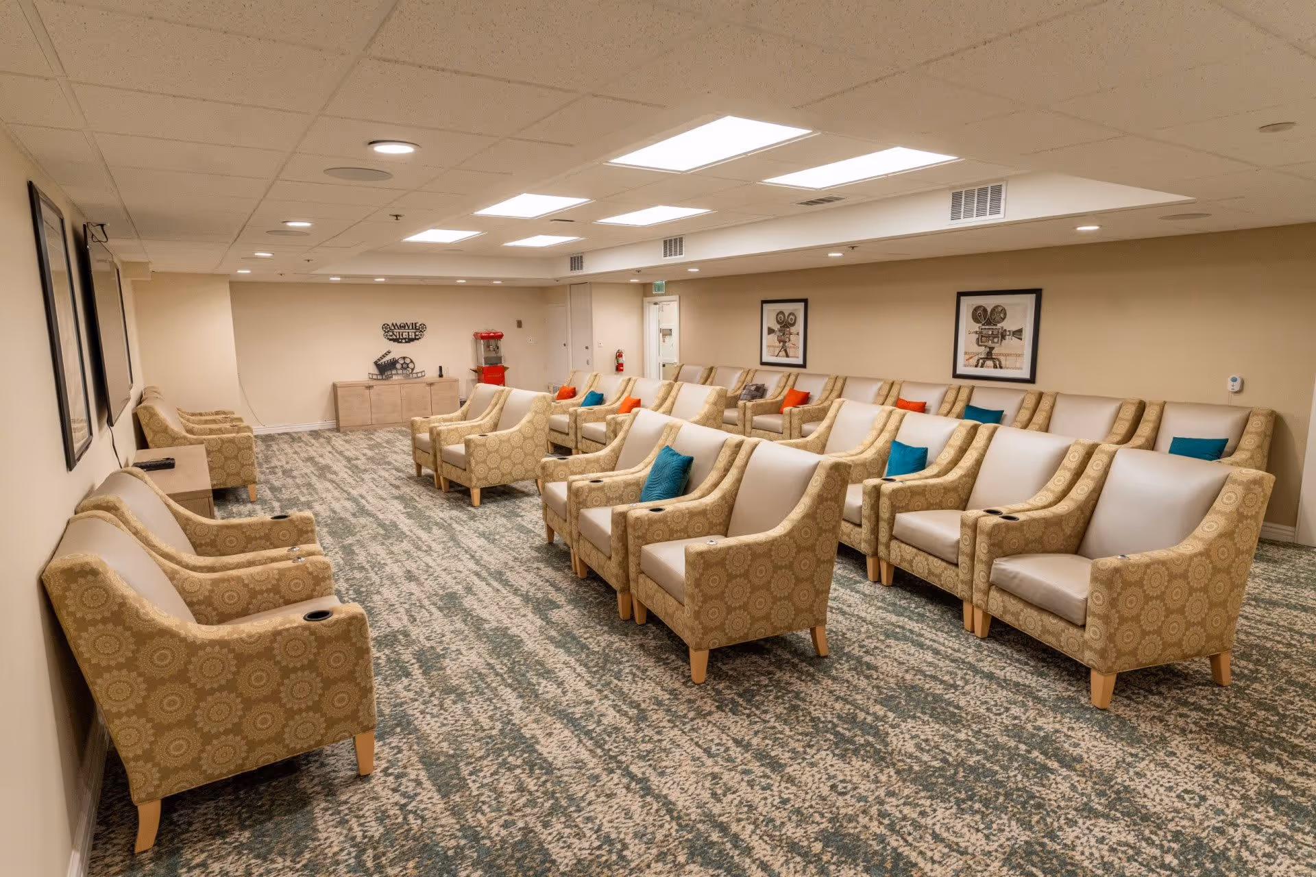 A cozy movie theater room with multiple rows of beige armchairs, each with a patterned fabric and leather seat cushions. Some chairs have colorful throw pillows in blue, red, and orange. The room has a carpeted floor with a green and beige pattern, beige walls, and framed movie-themed artwork. There is a popcorn machine and a cabinet at the far end of the room.