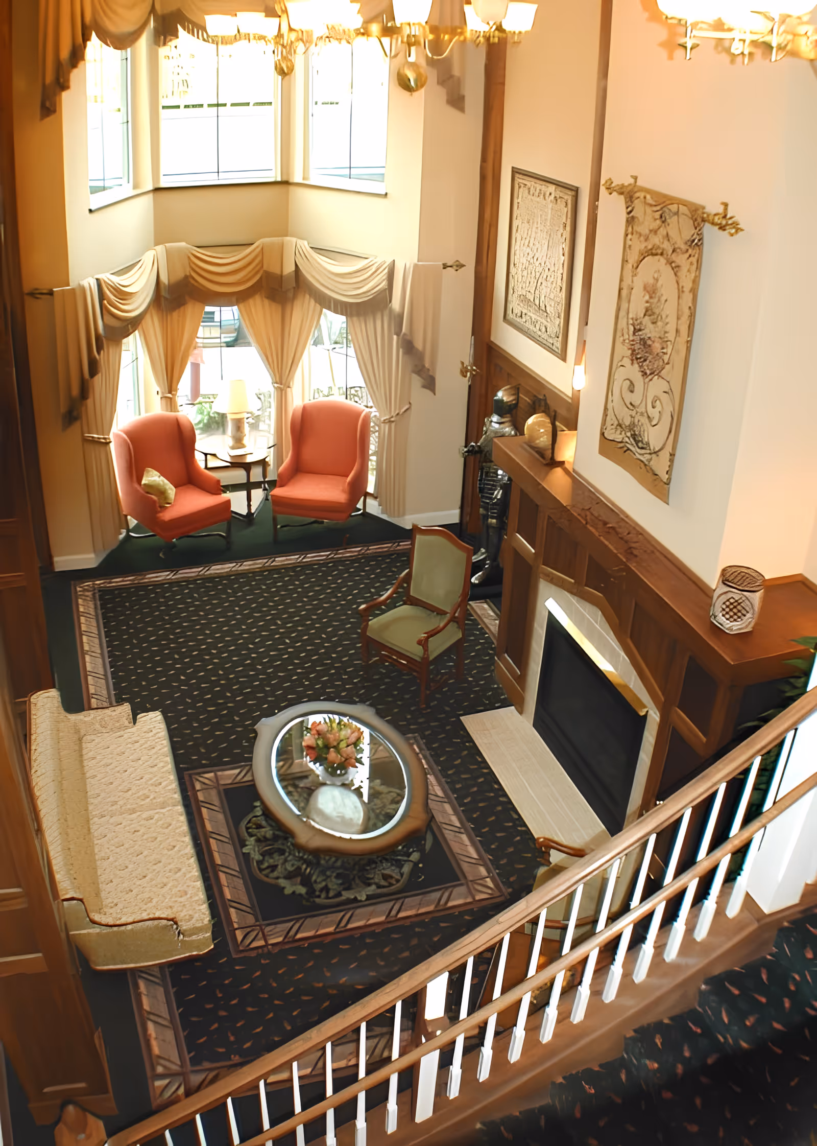 A cozy living room area viewed from above, featuring a fireplace with wooden mantel, two coral armchairs near a window with draped curtains, a beige sofa, a green upholstered chair, and a glass coffee table with a floral arrangement. The room has a patterned carpet and decorative wall hangings.