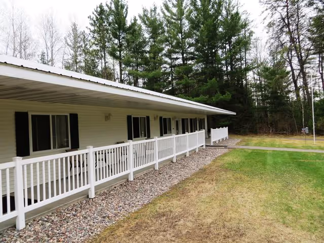 Exterior view of a single-story building with a white railing along a covered porch. The building has multiple windows with black shutters and is surrounded by grass and trees in the background.
