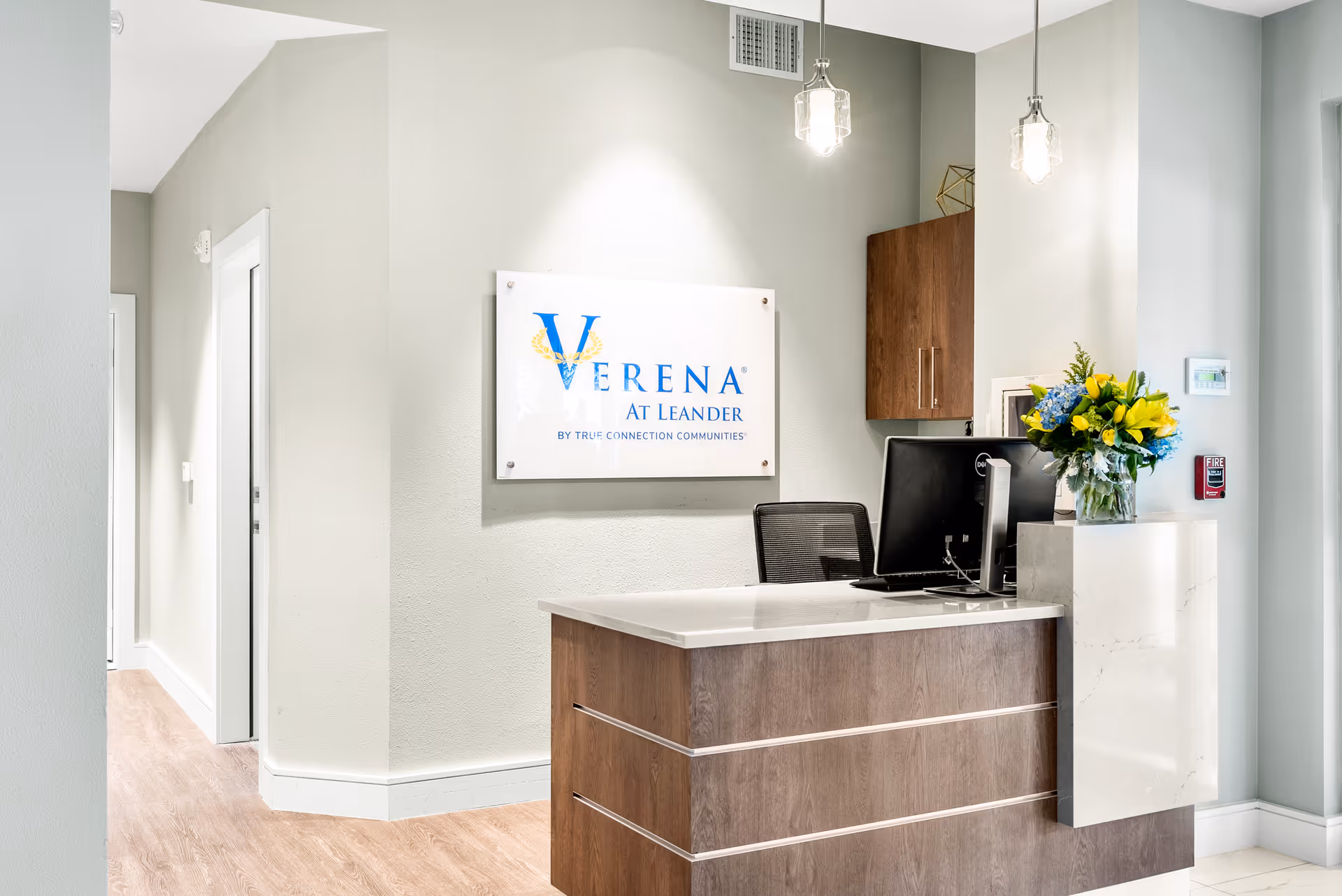 Reception desk area at Verena at Leander facility with a computer, a vase of yellow and blue flowers, wooden cabinets, and a sign on the wall displaying the facility name.