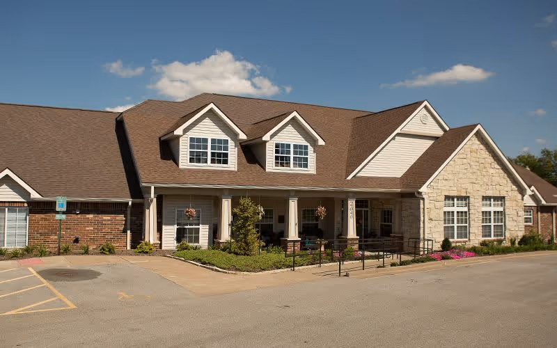 Exterior view of a single-story senior living facility building with a combination of brick and stone facade, multiple windows, a covered entrance with columns, and a wheelchair accessible ramp. The sky is clear with a few clouds.