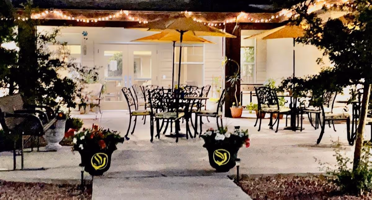Outdoor patio area at night with metal tables and chairs, yellow umbrellas, string lights overhead, potted plants, and a pathway leading to glass doors of a building.