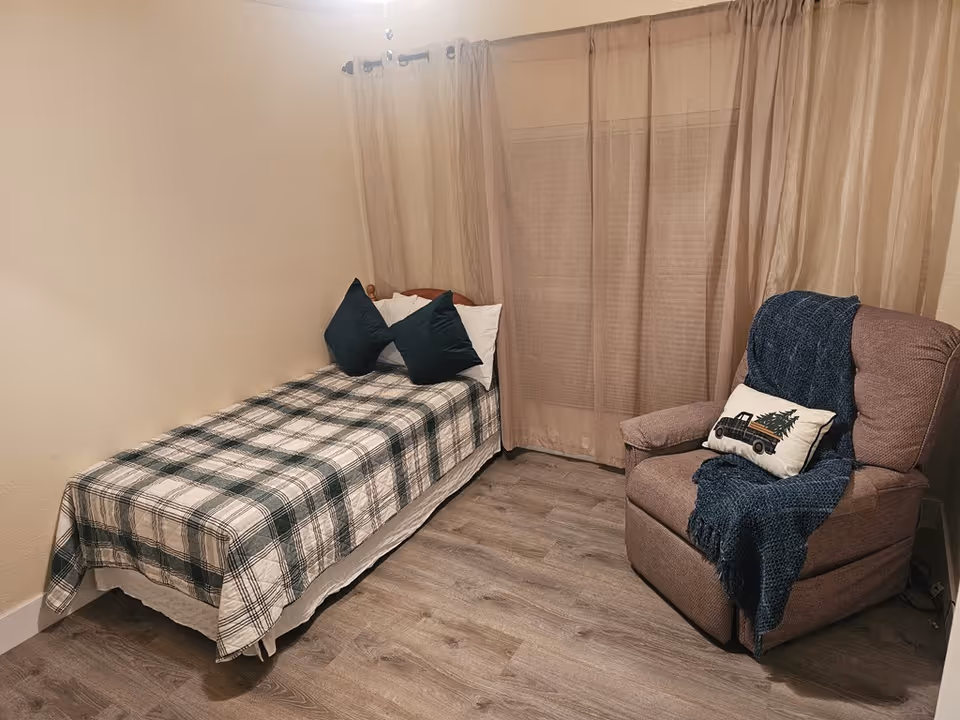 A small bedroom with a single bed covered in a plaid blanket and two dark green pillows. Next to the bed is a cushioned armchair with a blue throw blanket and a decorative pillow featuring a truck and tree design. Beige curtains cover the window behind the chair, and the floor is a light wood laminate.