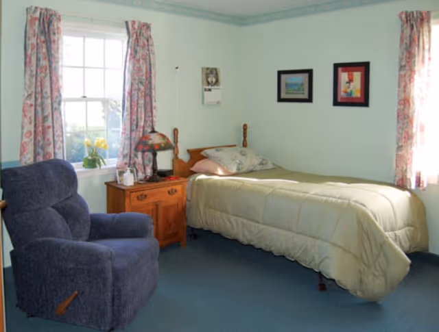 A small furnished bedroom with a twin bed, wooden nightstand, floral curtains at a window, and a blue recliner chair.