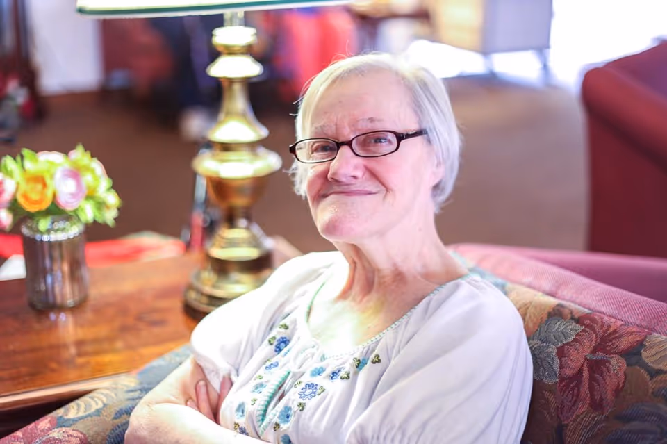 An elderly woman with short white hair and glasses sitting on a floral-patterned couch in a warmly lit living room, smiling gently with her arms crossed. A brass lamp and a small vase of flowers are visible on a wooden table beside her.