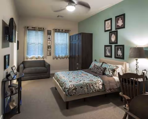 Bright senior living bedroom with a bed, wardrobe, seating area, and framed wall photos.