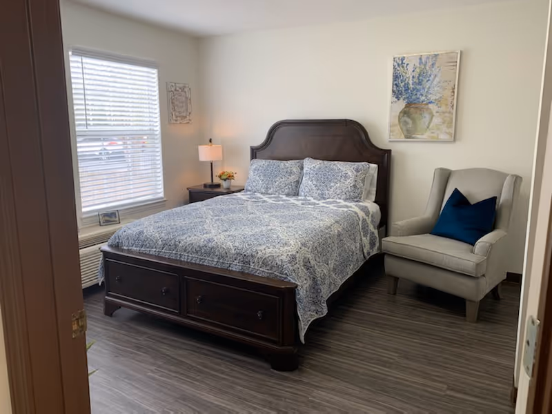 A bedroom with a wooden bed frame and a blue patterned bedspread. There are two matching pillows on the bed. To the left of the bed is a nightstand with a lamp and a small flower arrangement. A window with white blinds is on the left wall. On the right side of the bed is a gray armchair with a dark blue pillow. A painting of a vase with blue flowers hangs on the wall above the chair. The floor is a dark wood laminate.