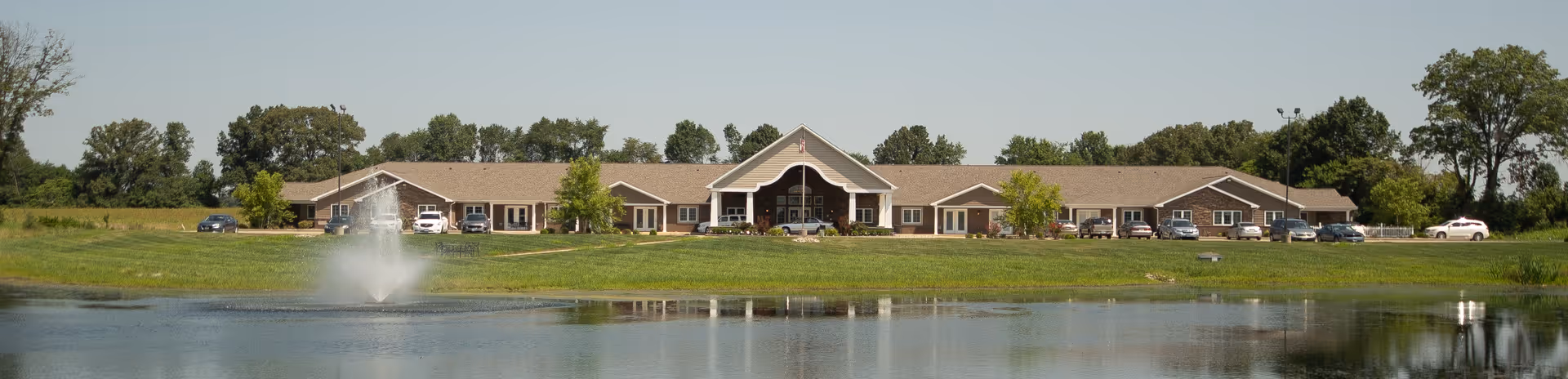 Wide exterior view of Villas of Holly Brook Herrin facility with a large pond and fountain in the foreground, surrounded by green grass and trees under a clear sky.