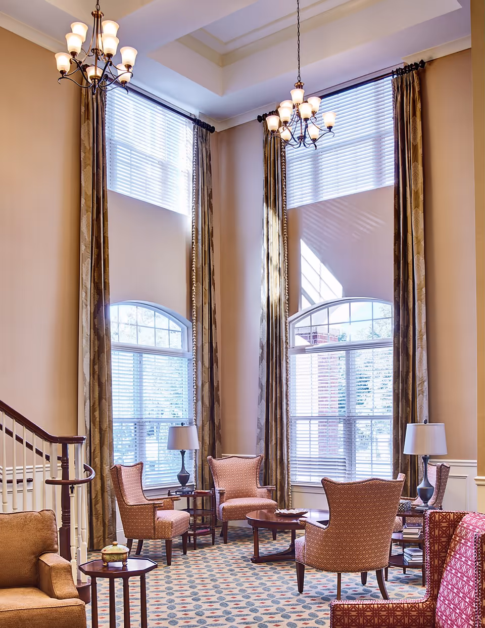 A bright and elegant living room area with tall windows covered by blinds and long patterned curtains. The room features several upholstered armchairs arranged around wooden coffee tables, two table lamps, and a patterned carpet. A staircase with a wooden handrail is visible on the left side.