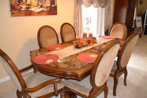 A dining room with a wooden dining table set for six people. The table has a decorative runner with candles and autumn-themed decorations in the center. There are six upholstered chairs around the table. A painting hangs on the wall and a window with draped curtains is visible in the background.