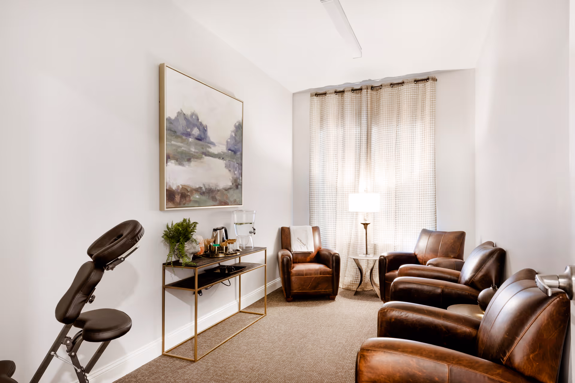 A cozy room with four brown leather armchairs arranged around a small round table with a lamp. A console table against the wall holds a water dispenser, coffee pot, cups, and a small plant. A large abstract painting hangs above the console table. The room has beige carpet and light-colored walls with a window covered by light curtains.