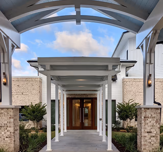 Covered walkway with white wooden beams leading to a building entrance with double wooden doors, surrounded by brick and white siding walls, with small landscaped bushes on either side and a partly cloudy blue sky overhead.